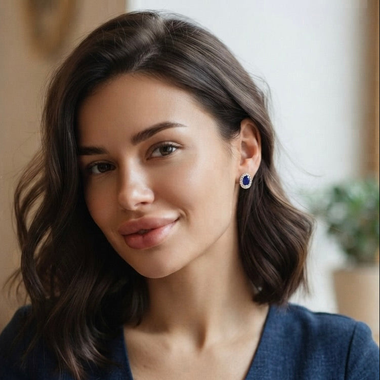A woman with wavy brown hair wearing a small blue oval shape sapphire stud earring, shown in a close-up portrait against a softly blurred indoor background.