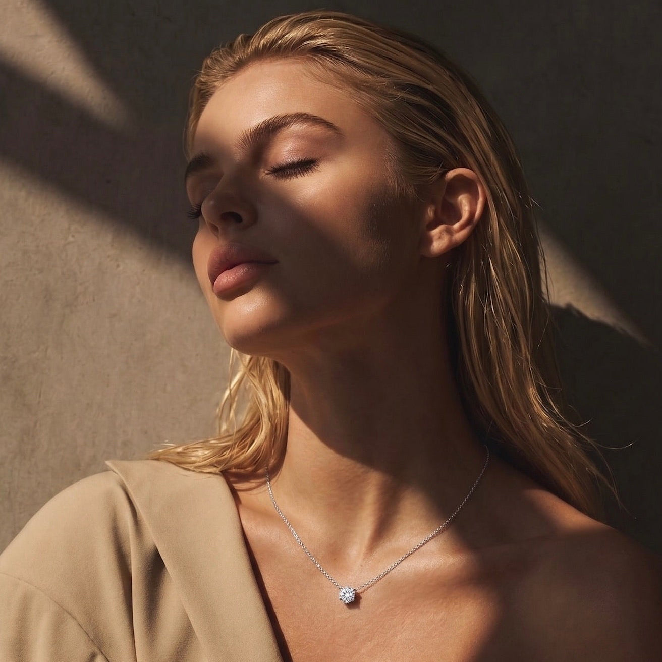 A woman with closed eyes wearing a delicate white gold necklace with a small round clear diamond, styled with a beige blazer against a textured wall with dramatic shadows.