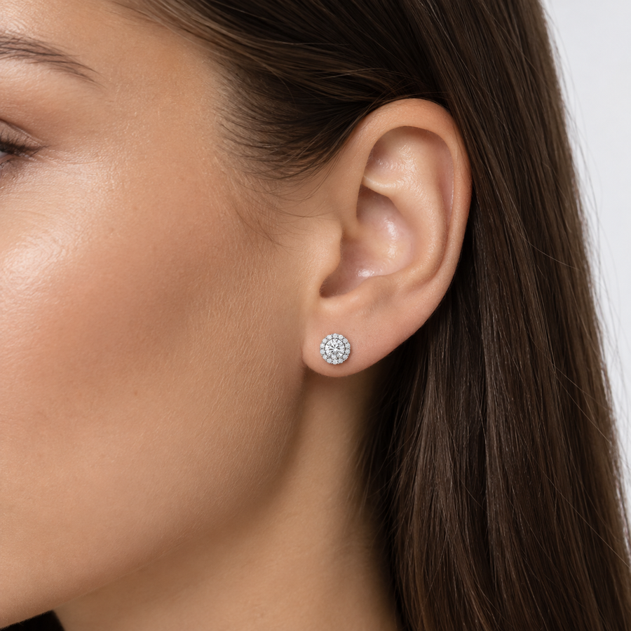 A close-up of a woman's ear wearing a small round stud earring with a halo of clear stones in white gold setting.