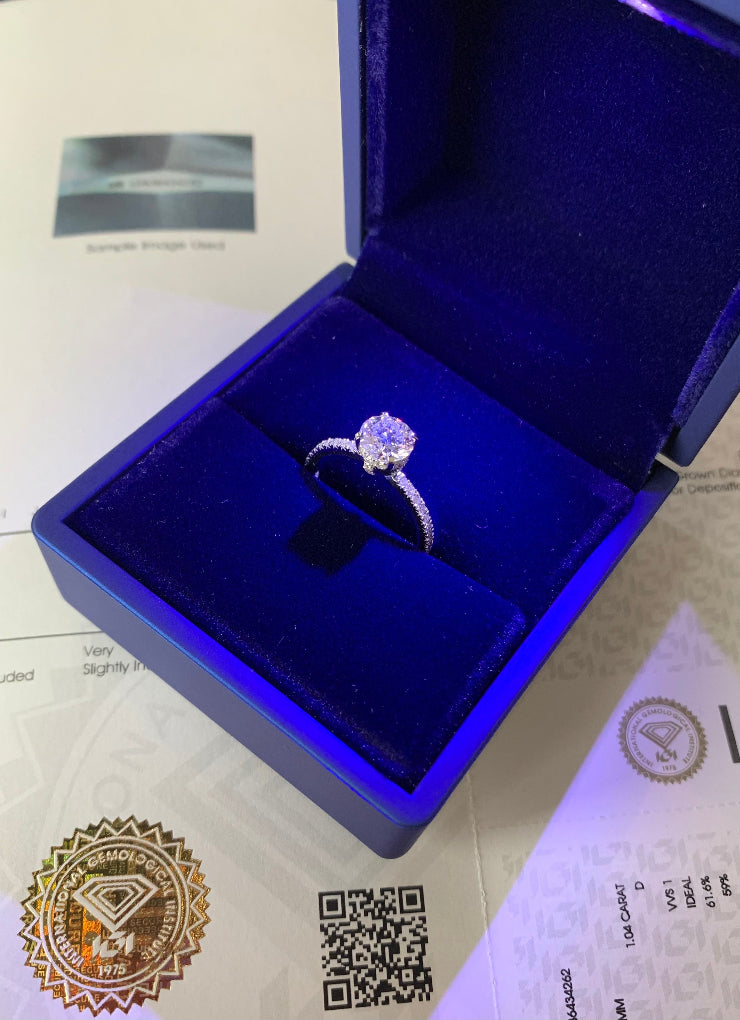 A white gold diamond engagement ring displayed in an open dark blue velvet ring box, with certification papers visible underneath.