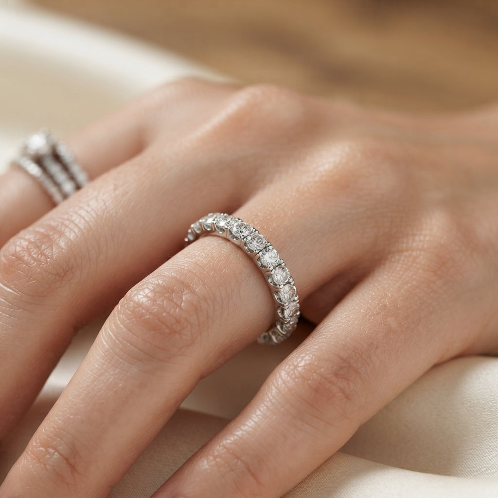 A close-up of a hand wearing a gold  diamond eternity band ring, with a second ring partially visible on the adjacent finger, set against a soft cream fabric background.
