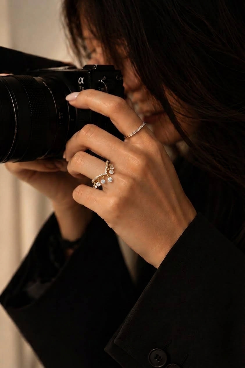 A close-up lifestyle shot of a woman holding a black camera, wearing a gold diamond ring with a floral cluster design on her finger.