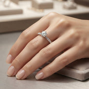 A close-up of a hand wearing a gold diamond engagement ring with a round center stone and a pavé band, resting on a neutral jewelry display surface.