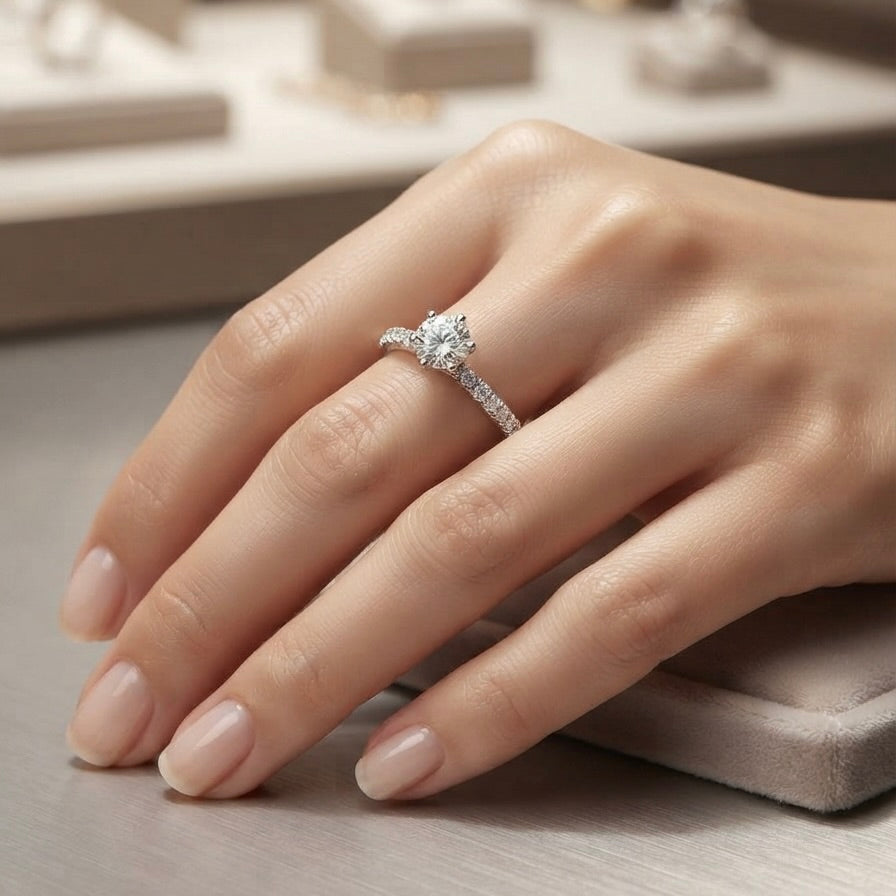 A close-up of a hand wearing a gold diamond engagement ring with a round center stone and a pavé band, resting on a neutral jewelry display surface.
